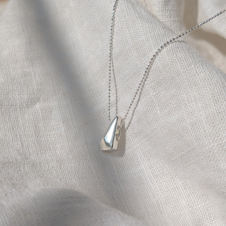 The Forbidden Facets Pendant by Becca Calmont, handmade from recycled sterling silver, features a smooth teardrop charm and is shown resting on textured white fabric in bright natural light.