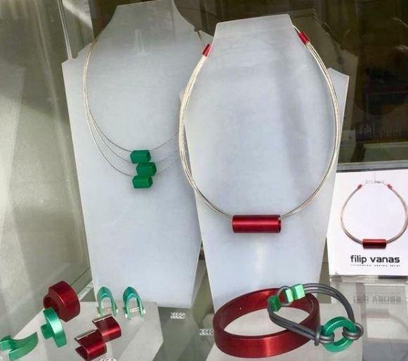 A jewelry display showcasing modern, minimalist pieces: two Tensor Tube Pendants by Filip Vanas with cylindrical anodised aluminium pendants (one red, one green), one red bracelet, a set of green earrings, and a grey bracelet with green accents. The designer's name, "filip vanas," is visible on a small card.