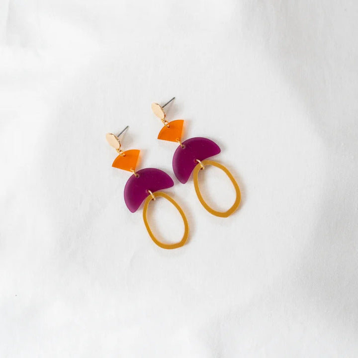 A pair of geometric statement earrings with vibrant colors crafted from 100% recycled acrylic. Each Magenta & Orange Fire Acrylic Earring by Studio Nok Nok has a metallic post with an orange triangle, a purple semicircle, and a mustard-yellow oval. They are displayed against a white fabric background.