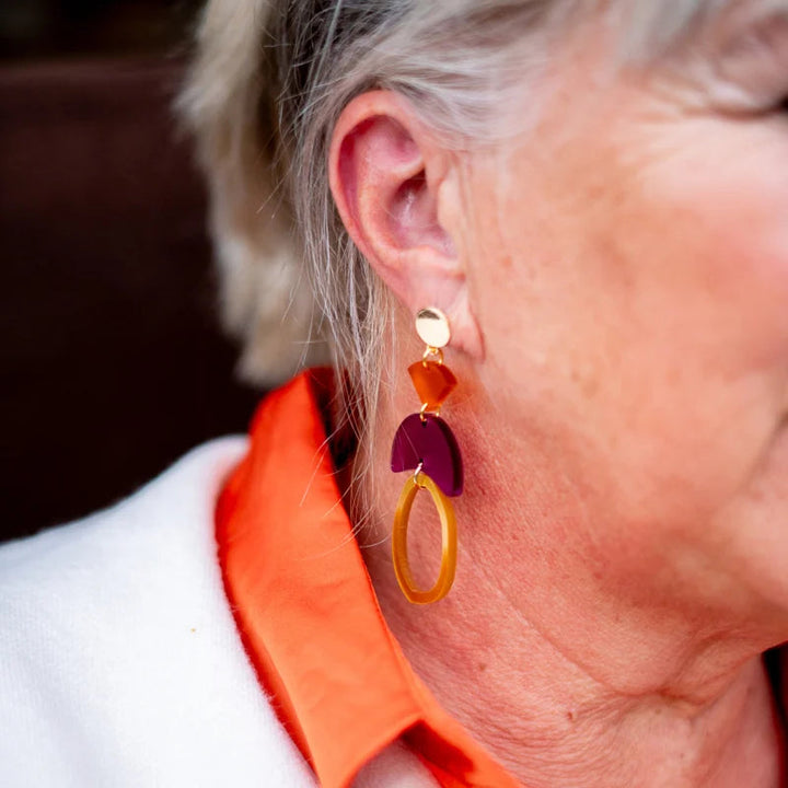 Close-up image of a person wearing Magenta & Orange Fire Acrylic Earrings by Studio Nok Nok, composed of yellow, pink, and purple geometric shapes made from 100% recycled acrylic. The person has light skin and is wearing an orange collared shirt with a white garment over it. Only part of their face, ear, and hair is visible.