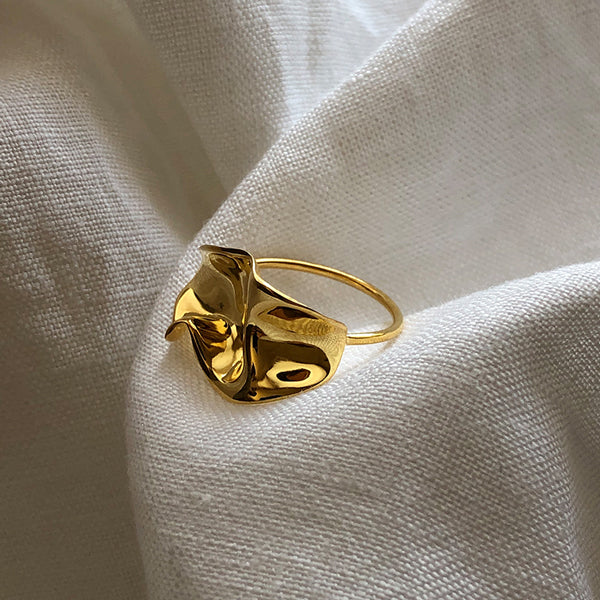 A handmade Flor Ring with an abstract, sculpted design by Bonvo is displayed on a textured white fabric backdrop. The 18ct gold plating highlights the smooth, flowing contours that catch the light, creating a sophisticated and elegant appearance.