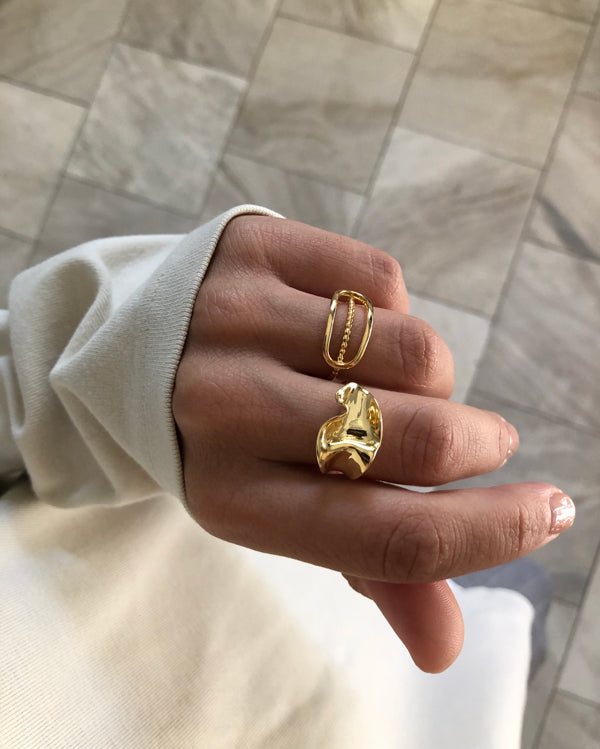 A person with a light skin tone, wearing a white long-sleeved shirt, displays two handmade rings on their fingers. One ring has a rectangular shape with a chain-like design, crafted from solid 925 sterling silver by Bonvo, while the other features a bold, sculpted form with 18ct gold plating from their Flor Ring collection. They are in a neutral-tone room with tiled flooring.