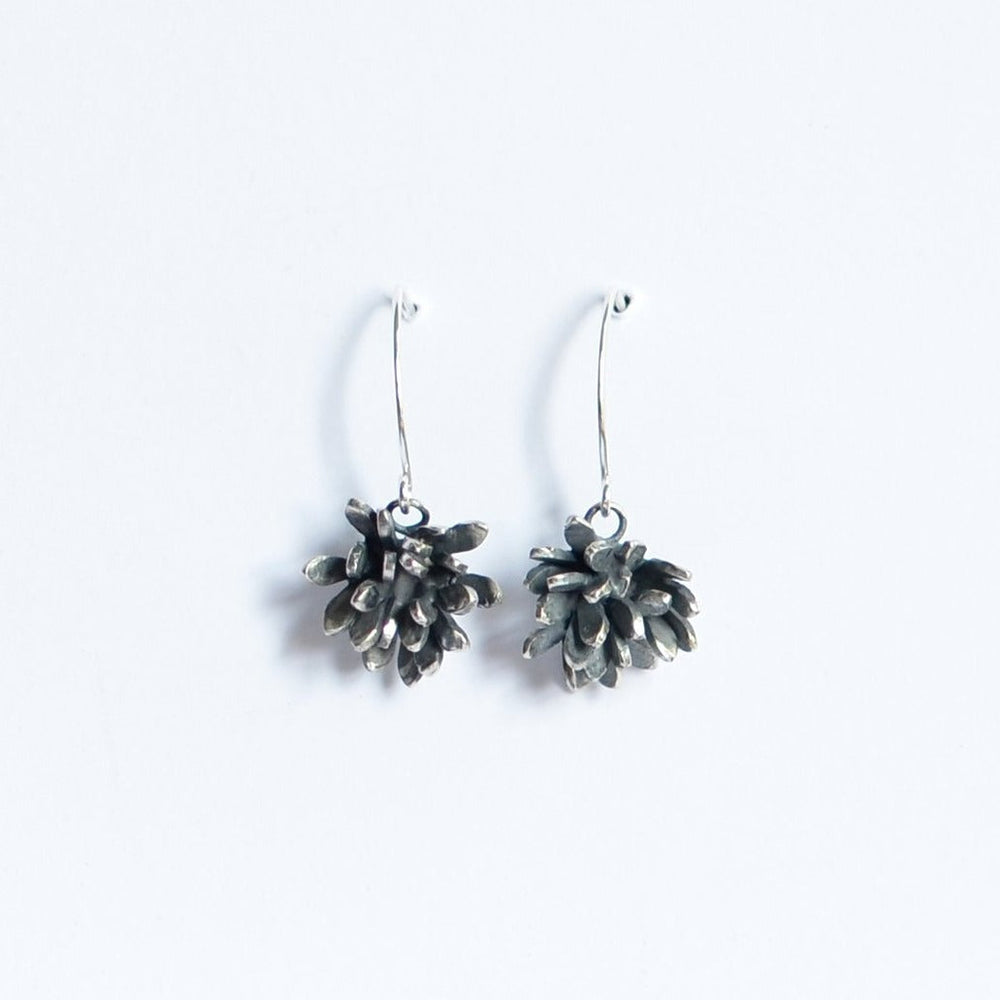 A pair of Folia Oxidised Silver Drop Earrings by Abbie Dixon is displayed against a plain white background. Each earring features a cluster of oxidised petals in a flower-like design, hanging from a curved wire hook.