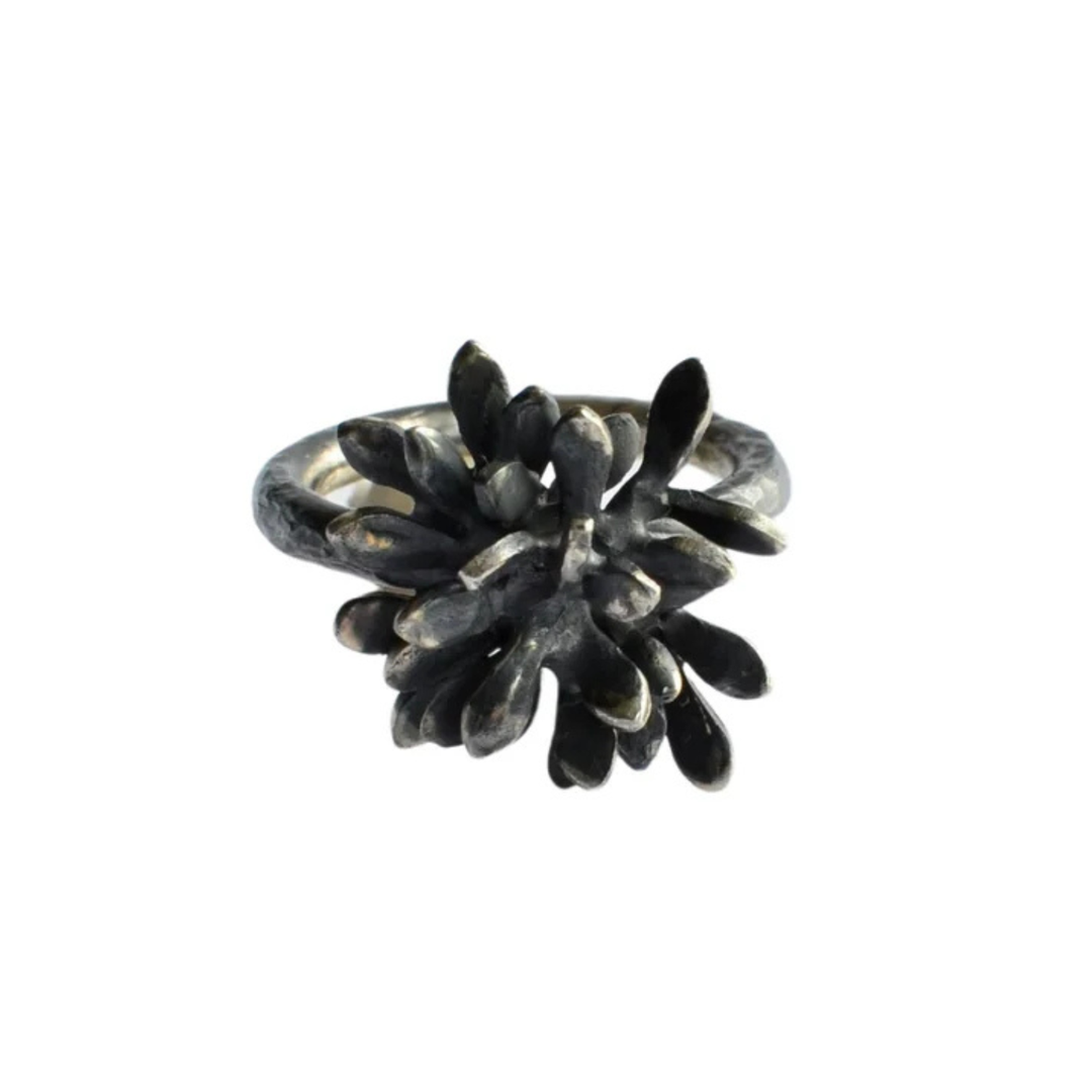 The Folia Oxidised Silver Ring by Abbie Dixon features an intricate, sculptural form with numerous small, petal-like elements radiating from the center, creating a textured, blossoming flower effect. The ring's darkened, oxidized Sterling silver finish adds an antique, artistic touch.