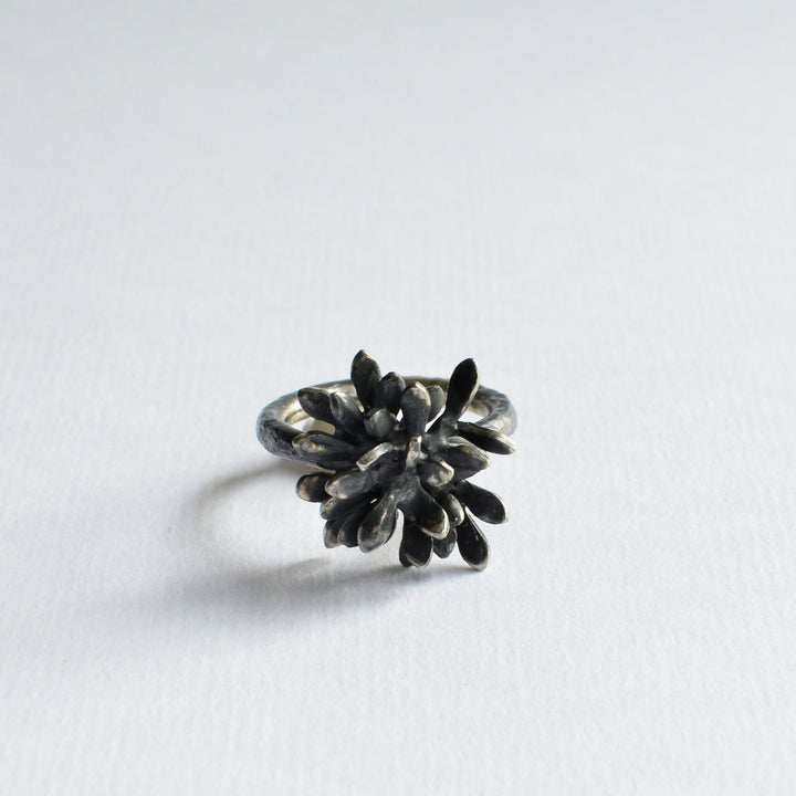 A handmade silver ring with a flower-shaped design on top, featuring multiple petals with a darkened, oxidized finish. The Folia Oxidised Silver Ring by Abbie Dixon is set against a plain white background.