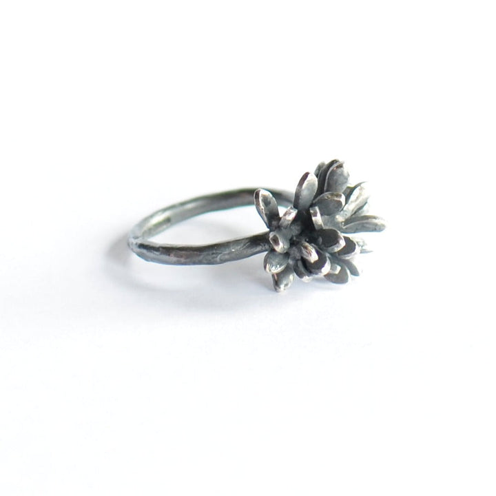 The Abbie Dixon Folia Oxidised Silver Ring features a detailed, sculpted flower with multiple petals. The flower design sits prominently on the thin, smooth band, creating an elegant and artistic piece of jewelry. The plain white background highlights the intricate design perfectly.