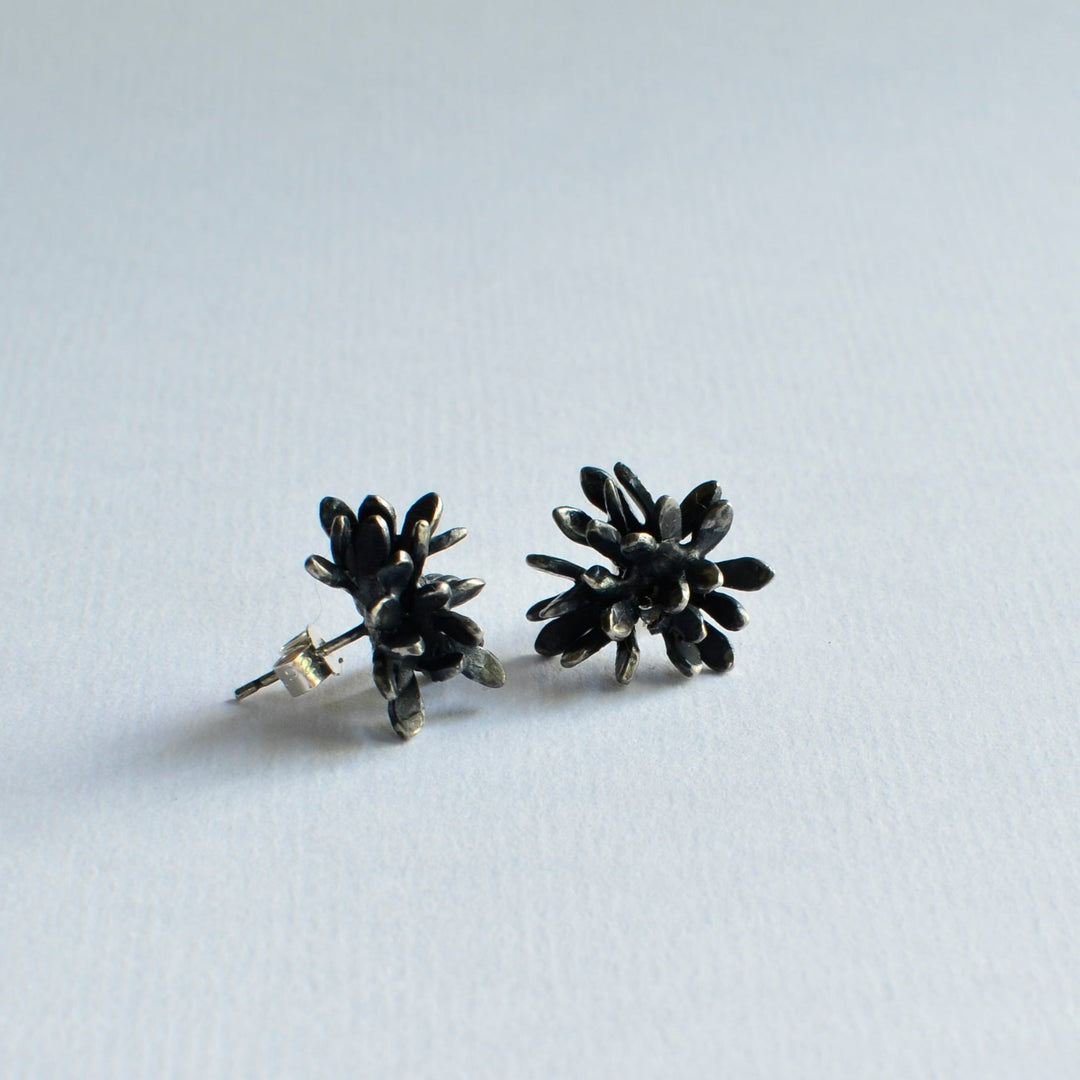 A pair of dark-toned, flower-shaped stud earrings crafted from oxidised sterling silver is displayed on a light background. The petals are clustered together, creating a textured, sculptural form. The earrings reflect subtle light, highlighting their metallic finish—introducing the Folia Oxidised Silver Studs by Abbie Dixon.