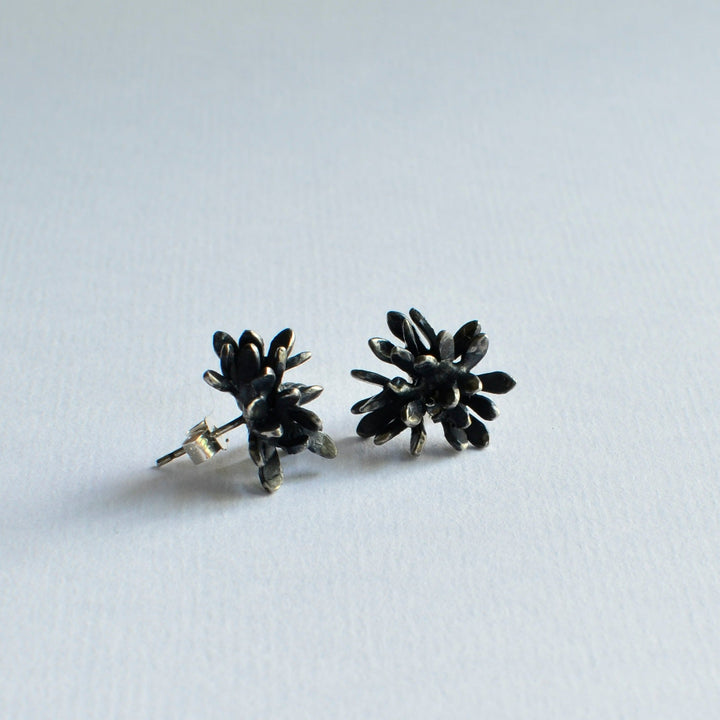 A pair of dark-toned, flower-shaped stud earrings crafted from oxidised sterling silver is displayed on a light background. The petals are clustered together, creating a textured, sculptural form. The earrings reflect subtle light, highlighting their metallic finish—introducing the Folia Oxidised Silver Studs by Abbie Dixon.