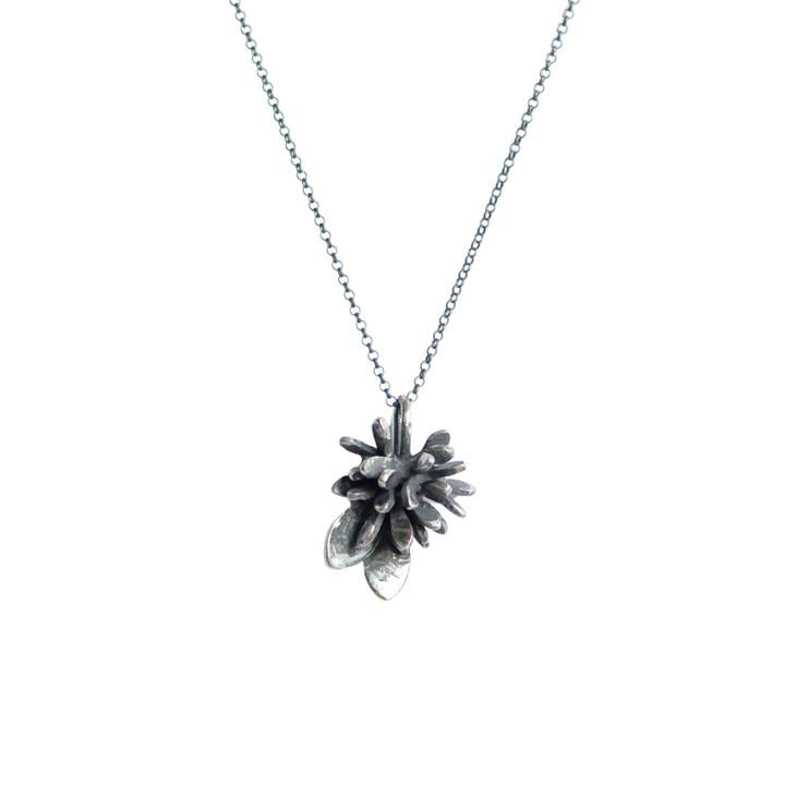 A delicate silver necklace featuring a pendant shaped like a small pinecone. The Folia Oxidised Silver Pendant, crafted by Abbie Dixon in oxidised sterling silver, hangs from a fine chain, showcasing intricate details that resemble the natural form of a pinecone with overlapping scales. The background is white.