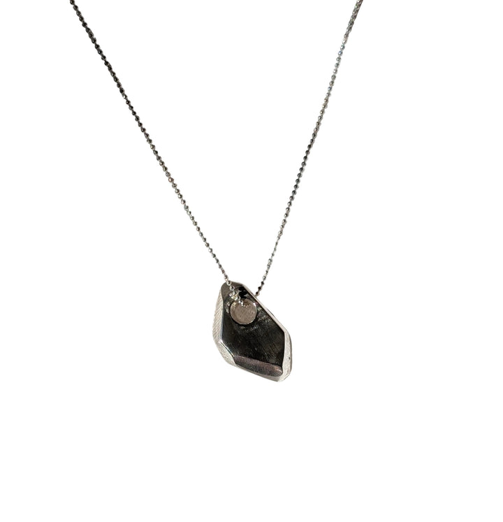 The Becca Calmont Forbidden Facets Pendant, a handmade sterling silver necklace with an irregular-shaped charm on an 18-inch chain, rests on softly wrinkled white fabric in natural sunlight with gentle shadows.