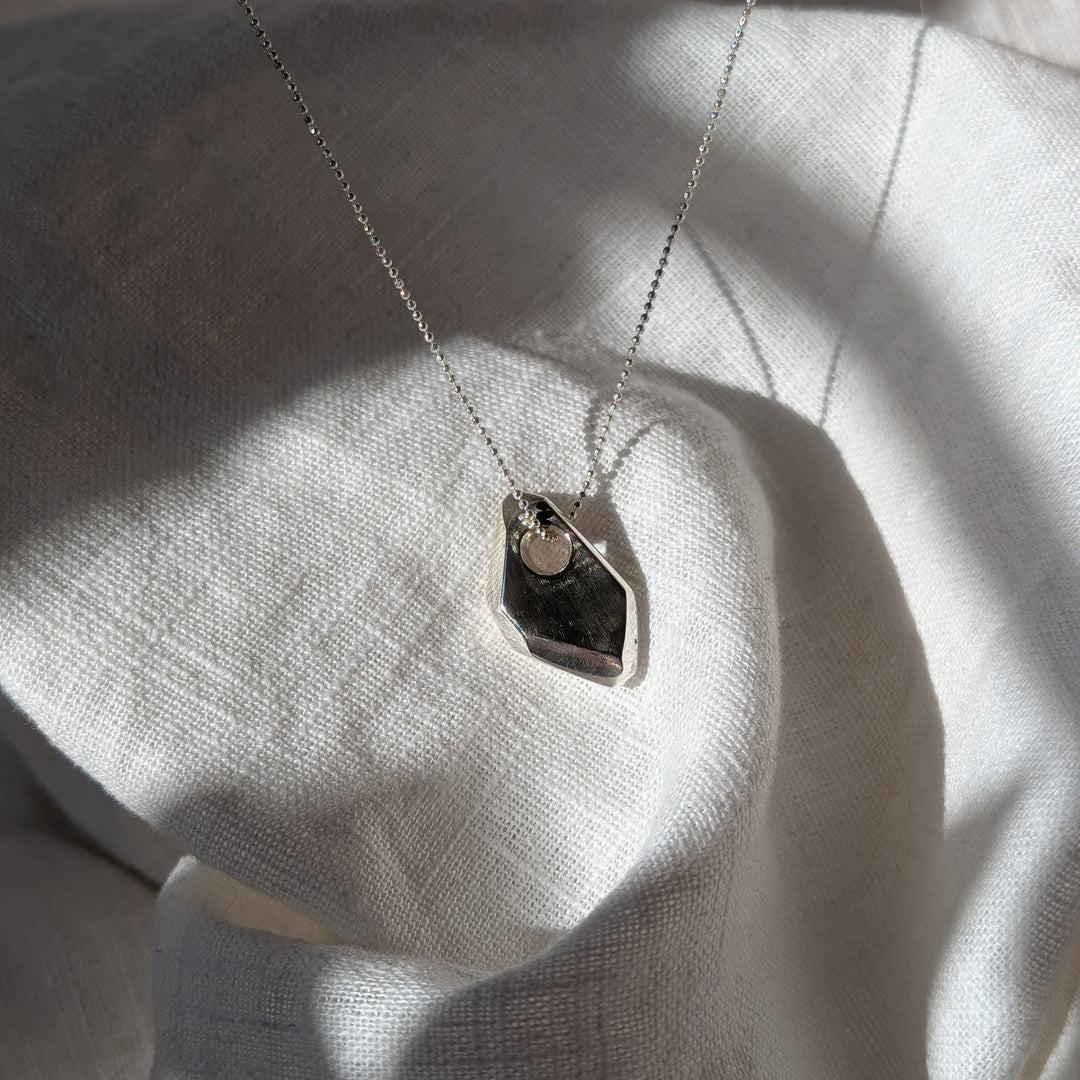 The Forbidden Facets Pendant by Becca Calmont, crafted from recycled sterling silver, lies on textured white fabric with sunlight accentuating its intricate design and elegant 18-inch chain.