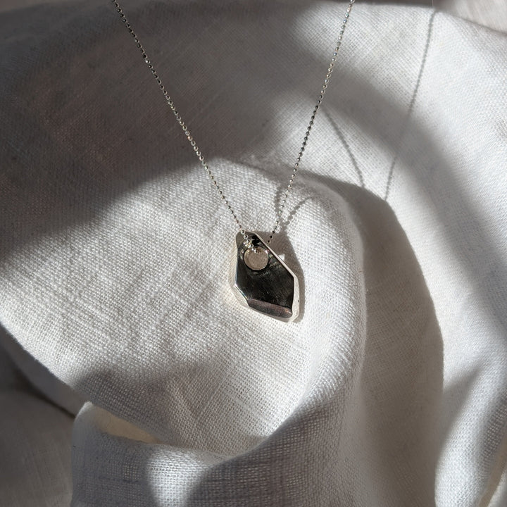 The Forbidden Facets Pendant by Becca Calmont, crafted from recycled sterling silver, lies on textured white fabric with sunlight accentuating its intricate design and elegant 18-inch chain.