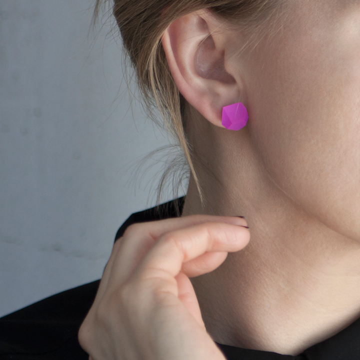 Close-up of a person wearing Fruit Bijoux Stud Earrings VU Crystals Magenta Pink (Silver or Gold). The vibrant geometric-shaped earrings are crafted from acrylic resin. The person has light skin and is touching their neck with their left hand. The background is a neutral shade, and a portion of the person's black clothing is visible.