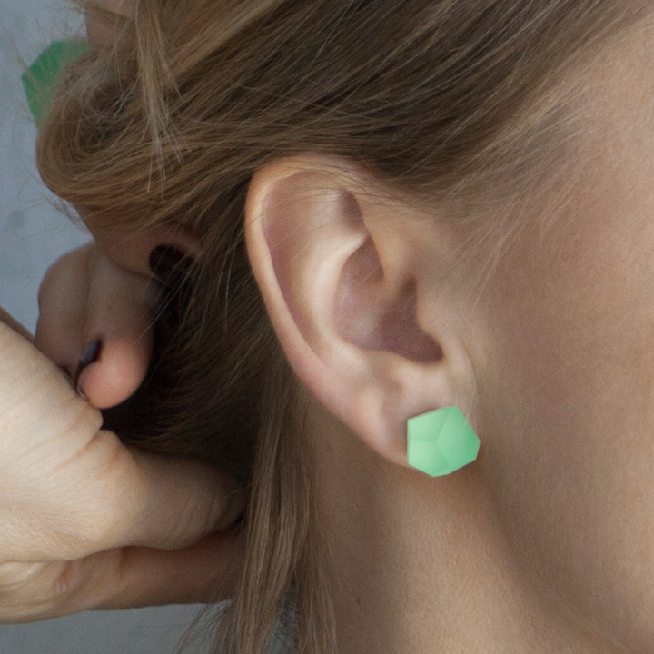 A close-up image of a person's ear adorned with the Fruit Bijoux Stud Earrings VU Crystals Flash Green in gold. The person's light brown hair is pulled back, and a hand with dark nail polish is seen adjusting the earring. The background is blurred.