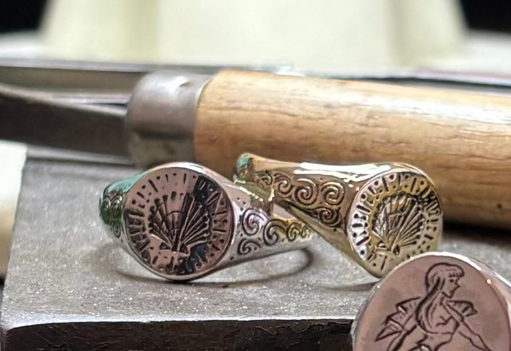 Three engraved silver rings rest on a jeweler’s workbench, including the Cosmic Boulevard Sterling Silver Siren Signet Ring, featuring hand-engraved details of a woman with fish tails and radiating patterns, set amidst tools in a blurred workshop.