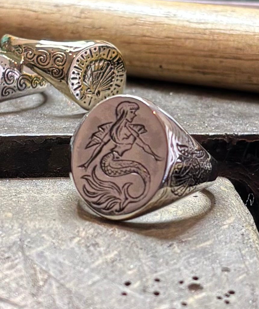 A Sterling Silver Siren Signet Ring by Cosmic Boulevard, featuring a hand-engraved mermaid design, rests on a workbench, while another ornate ring with a shell motif appears in the background.