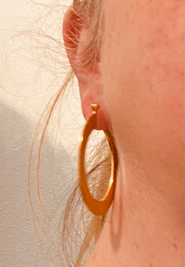 Close-up of a person wearing Gallardo & Blaine Designs Satin Hoop Earrings. The photograph shows part of the ear and the earring, with light hair strands visible against a light background. The medium-sized, oval-shaped earring gleams subtly.