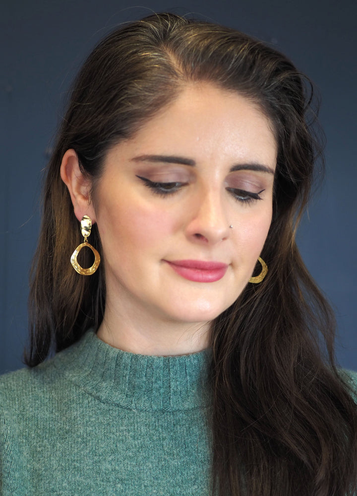These earrings are gold plated and have an elegant, organic design. The upper portion is an irregular, slightly concave oval with a textured, hammered finish that reflects light beautifully. Suspended from the top stud is a hollow, asymmetrical, open hoop with a wavy, freeform shape. The hoop also features a hammered texture with small, circular indentations, adding depth and visual interest. Pictured here on a model.