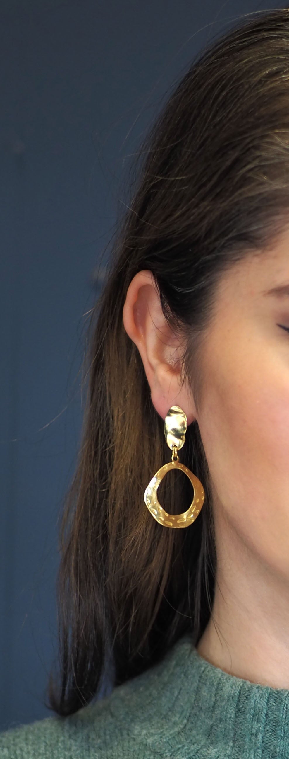 These earrings are gold plated and have an elegant, organic design. The upper portion is an irregular, slightly concave oval with a textured, hammered finish that reflects light beautifully. Suspended from the top stud is a hollow, asymmetrical, open hoop with a wavy, freeform shape. The hoop also features a hammered texture with small, circular indentations, adding depth and visual interest. Pictured here on a model.
