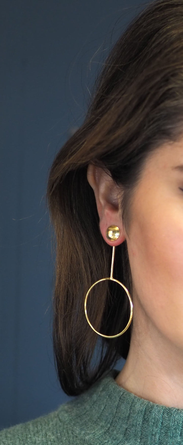 Close-up of a person wearing a long, elegant earring. The Watermelon tropical Lulu Gold Hoop Earrings feature a small stud on the earlobe from which a thin, vertical rod extends, ending in a large, dangling hoop. The person has medium-length dark hair and is wearing a green knit garment reminiscent of Sixties club fashion.