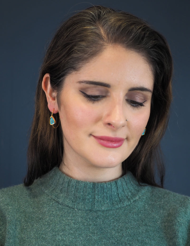 The Chloe Gold & Green Turquoise Hoop Earrings from Watermelon Tropical feature gold hoops with triangular blue Turquoise stones at the centre. The rustic appearance and dark striations of the stones enhance their natural beauty, while sterling silver prongs delicately secure them. These earrings boast a minimalist, elegant design that combines gold and black accents gracefully. Pictured here on a model.