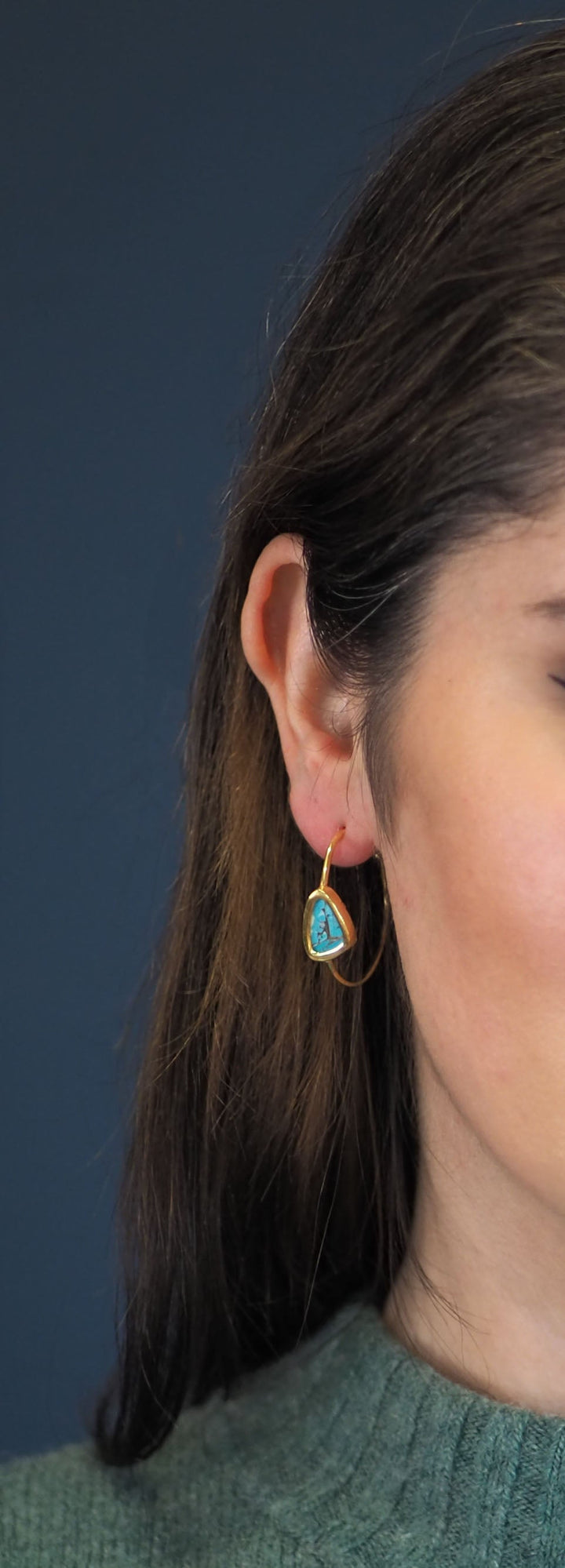 The Chloe Gold & Green Turquoise Hoop Earrings from Watermelon Tropical feature gold hoops with triangular blue Turquoise stones at the centre. The rustic appearance and dark striations of the stones enhance their natural beauty, while sterling silver prongs delicately secure them. These earrings boast a minimalist, elegant design that combines gold and black accents gracefully. Pictured here on a model.