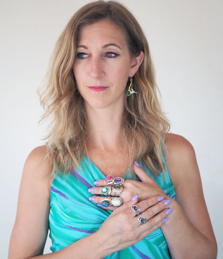 A woman with blonde hair is wearing a turquoise and blue dress. She has several colourful Eyetelia Rings in various gemstones by Gallardo & Blaine Designs on her fingers, their vibrant hues matched perfectly by her nail polish. Adorning her ear with a pendant earring, she glances thoughtfully to the side, making a striking statement.