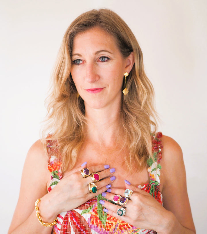 A woman with wavy blonde hair is wearing a colorful floral dress. Displaying an Art Deco Ring in Silver Gold & various gemstones by Gallardo & Blaine Designs and multiple rings with colorful gemstones on both hands, she accessorizes with a gold vermeil bracelet and geometric gold earrings. She poses against a plain white background.