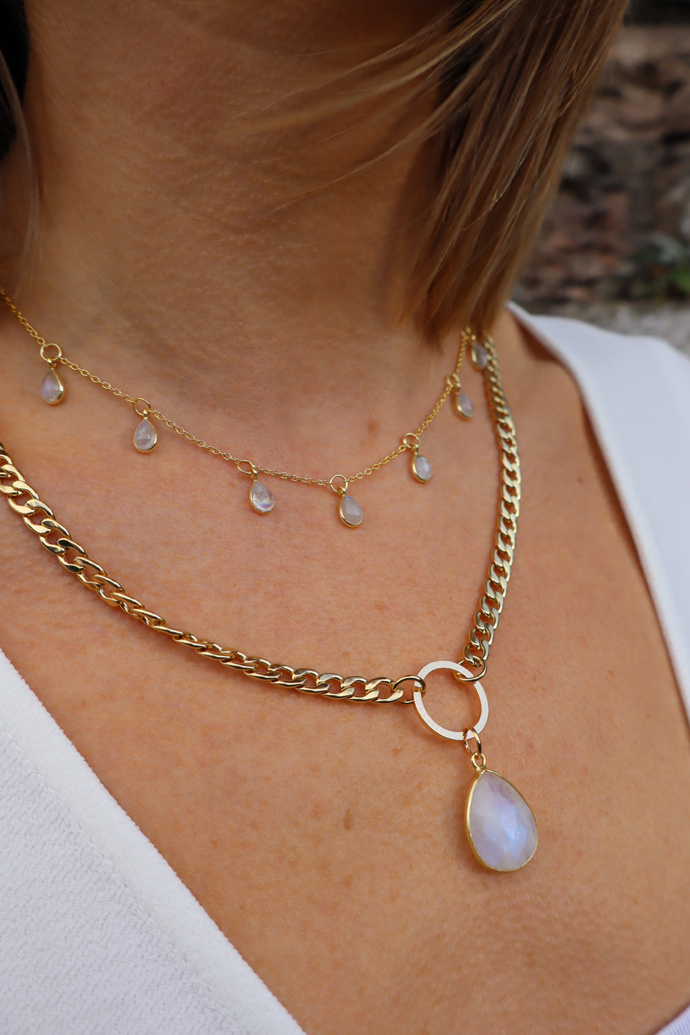 Close-up of a woman's neck wearing the Gaia Moonstone Necklace by Kybalion Jewellery, featuring small dangling pearls and a gold-plated curb chain with a large oval moonstone pendant, paired with a white top.