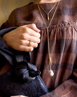 A person in a brown plaid dress holds a black textured knotted clutch. They wear a gold ring and layered gold necklaces, including Inner Island’s Gathered Forms Lariat Necklace crafted from recycled sterling silver.