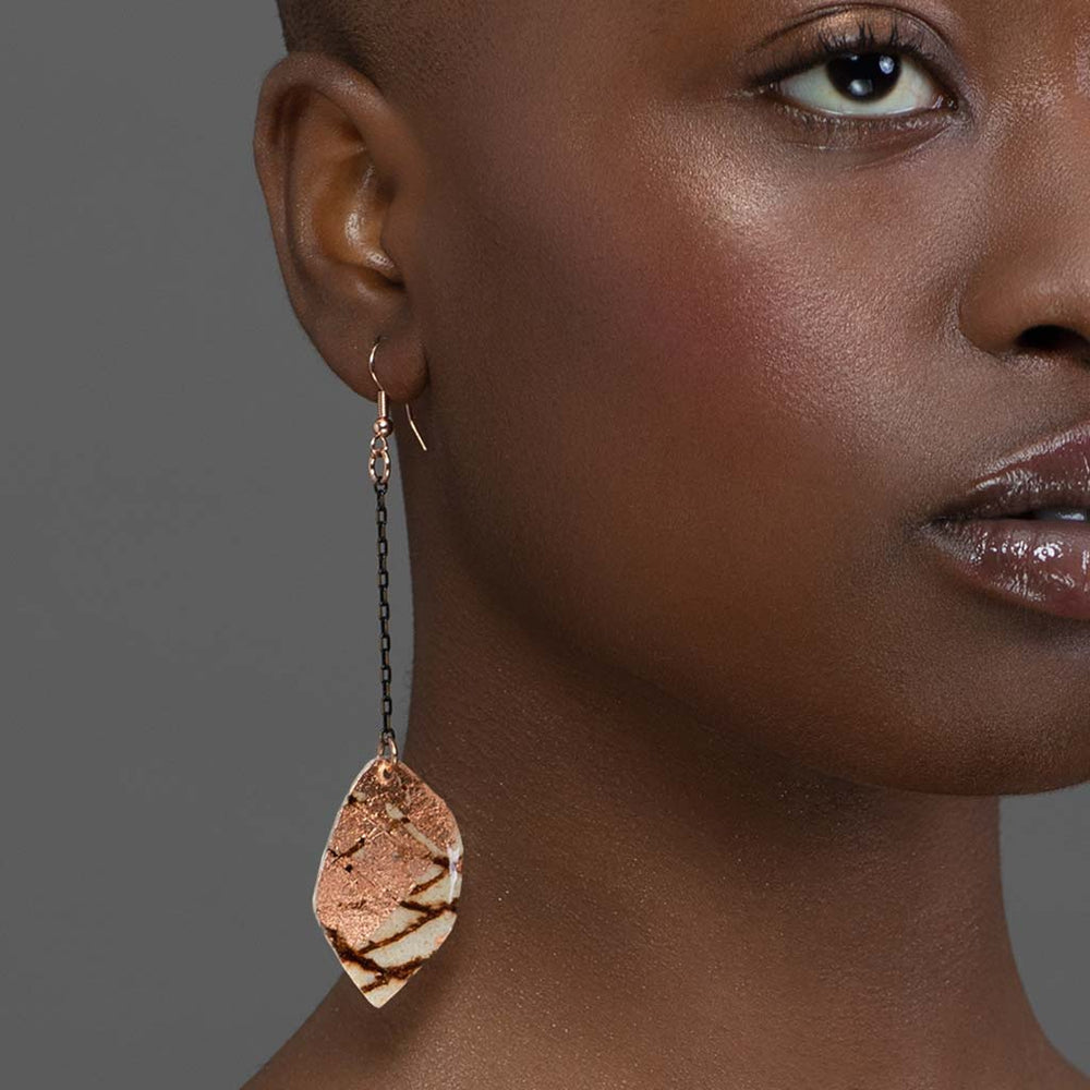 A close-up shows part of a dark-skinned person’s face and neck wearing Rothlú’s Gile Abstract Leaf Batik Textile Earrings in Rust/Rose Gold, featuring a unique handmade leaf-shaped pendant on a black chain against a grey background.
