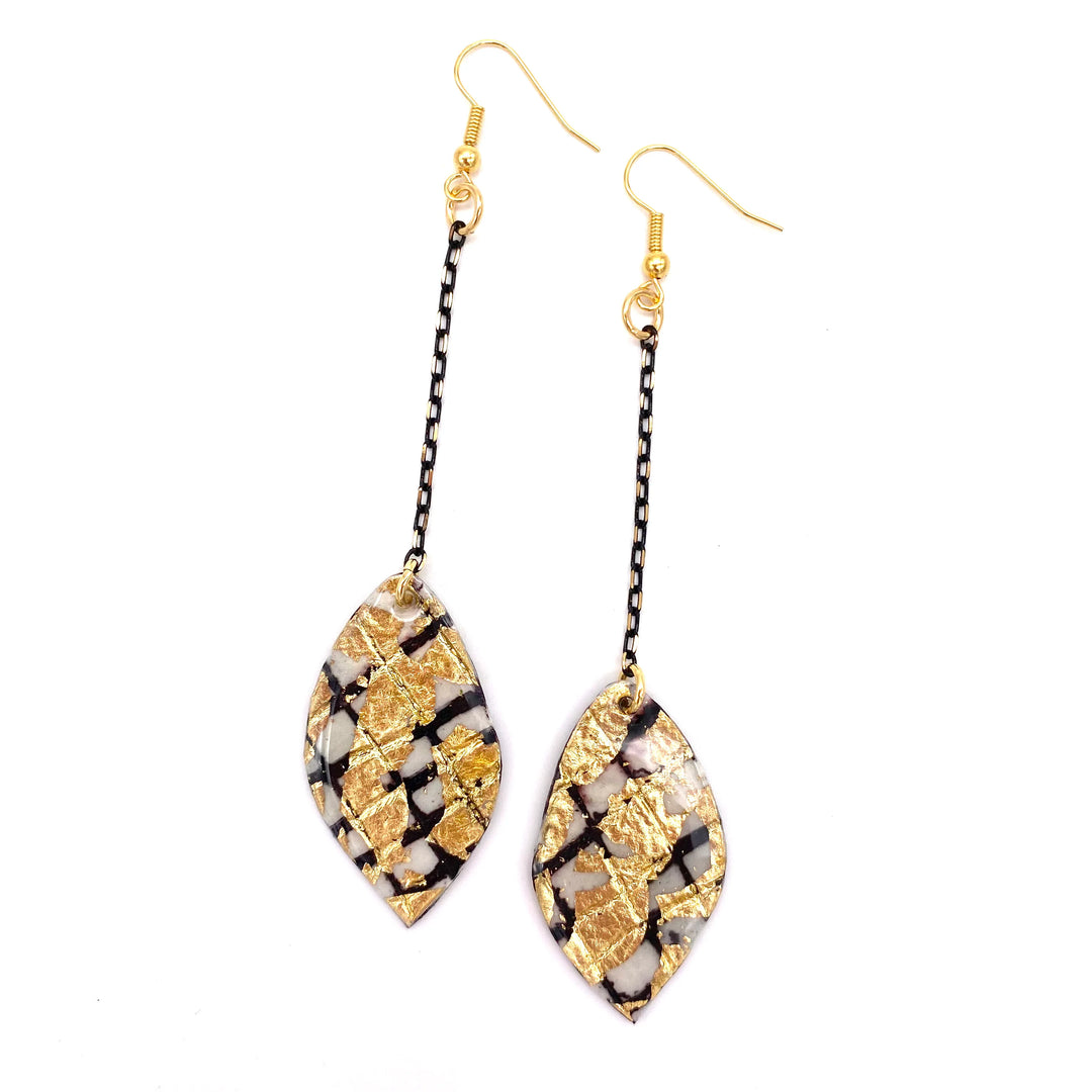 Introducing the Rothlú Gile Abstract Leaf Batik Textile Earrings in Black/Gold, a stunning pair of hypo-allergenic gold-plated earrings. These earrings feature leaf-shaped pendants adorned with a mosaic-like design in black, white, and gold. Each earring is elegantly connected to the pendant by a short black chain and finished with a gold hook.