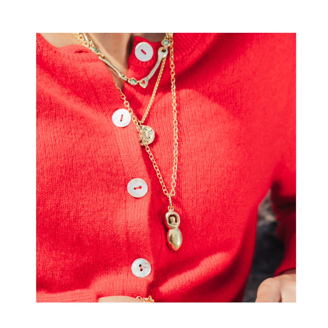 A person wearing a bright red buttoned cardigan with layered gold necklaces, one featuring the Goddess Pendant by Madeleine Sculptural Jewellery.