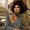 A woman with curly hair sits on outdoor metal stairs by a brick wall, wearing a patterned black and white outfit, layered necklaces, and the Elsiem Vibe Gold Dome Ring—a hand-painted statement piece—gazing thoughtfully into the distance.