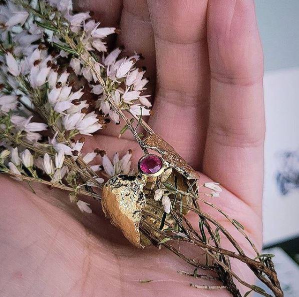 A hand holds an ethereal Faerie Tale Ring In Blue Topaz by Caroline Stokesberry-lee, with a blue topaz gemstone set in a rugged gold band, crafted using the reticulation technique and intertwined with small dry white flowers. The ring and flowers rest gently in the palm, showcasing intricate details of the jewelry and the delicate petals.