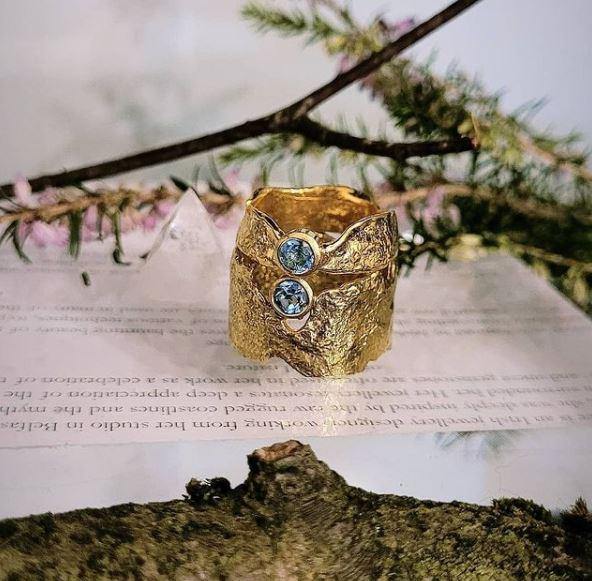 A rustic gold ring, crafted using the reticulation technique and adorned with blue topaz gemstones, is displayed on an open book. The Faerie Tale Ring In Blue Topaz by Caroline Stokesberry-lee is showcased beautifully. The background includes a floral arrangement with pink flowers and green foliage, adding a natural, organic feel to this ethereal faerie tale scene.