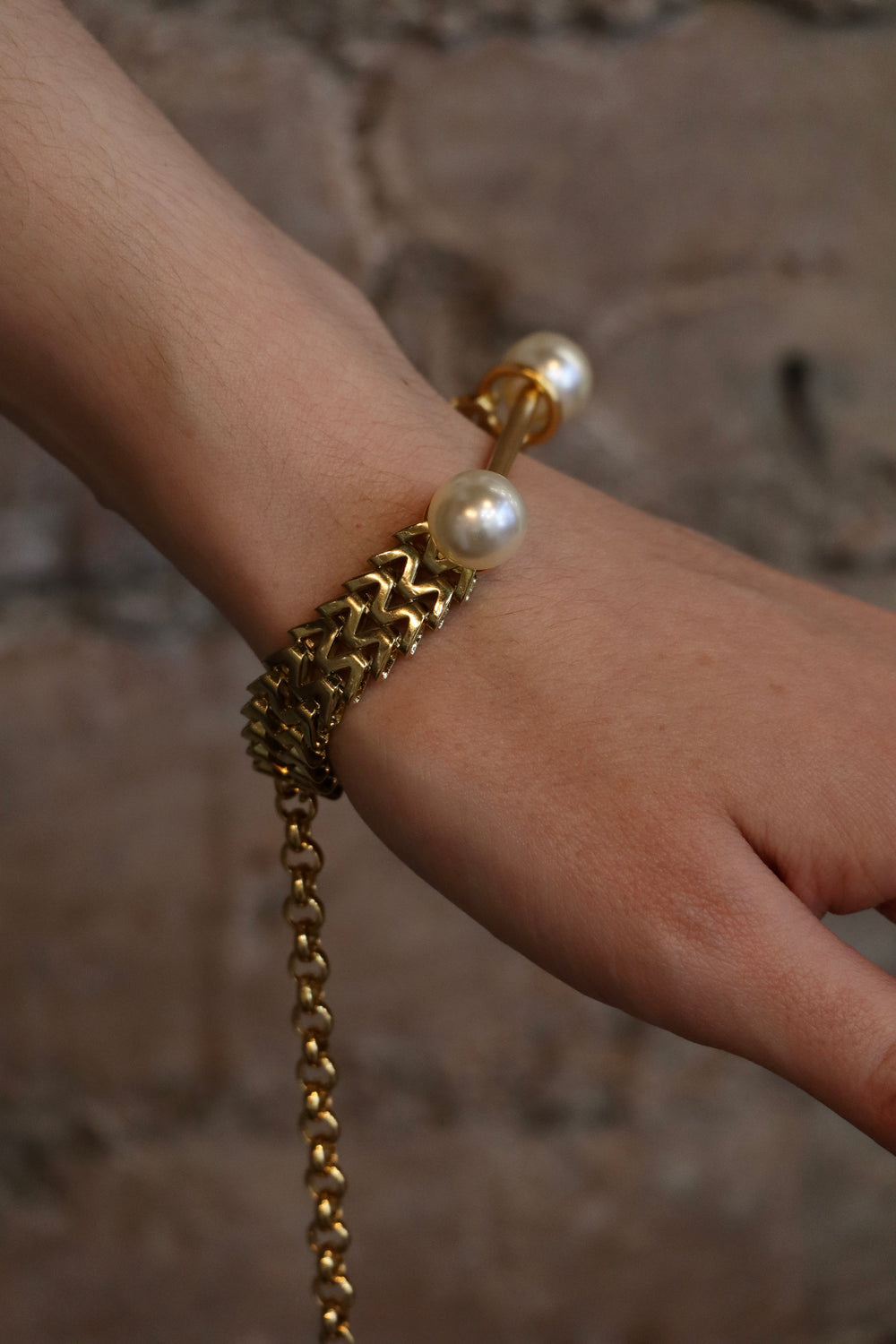 A close-up of a hand wearing the Zubeldia Gold Bar and Double Pearl Chunky Bracelet, featuring a chunky gold chain with two large pearls at each end, set against a blurred stone background.