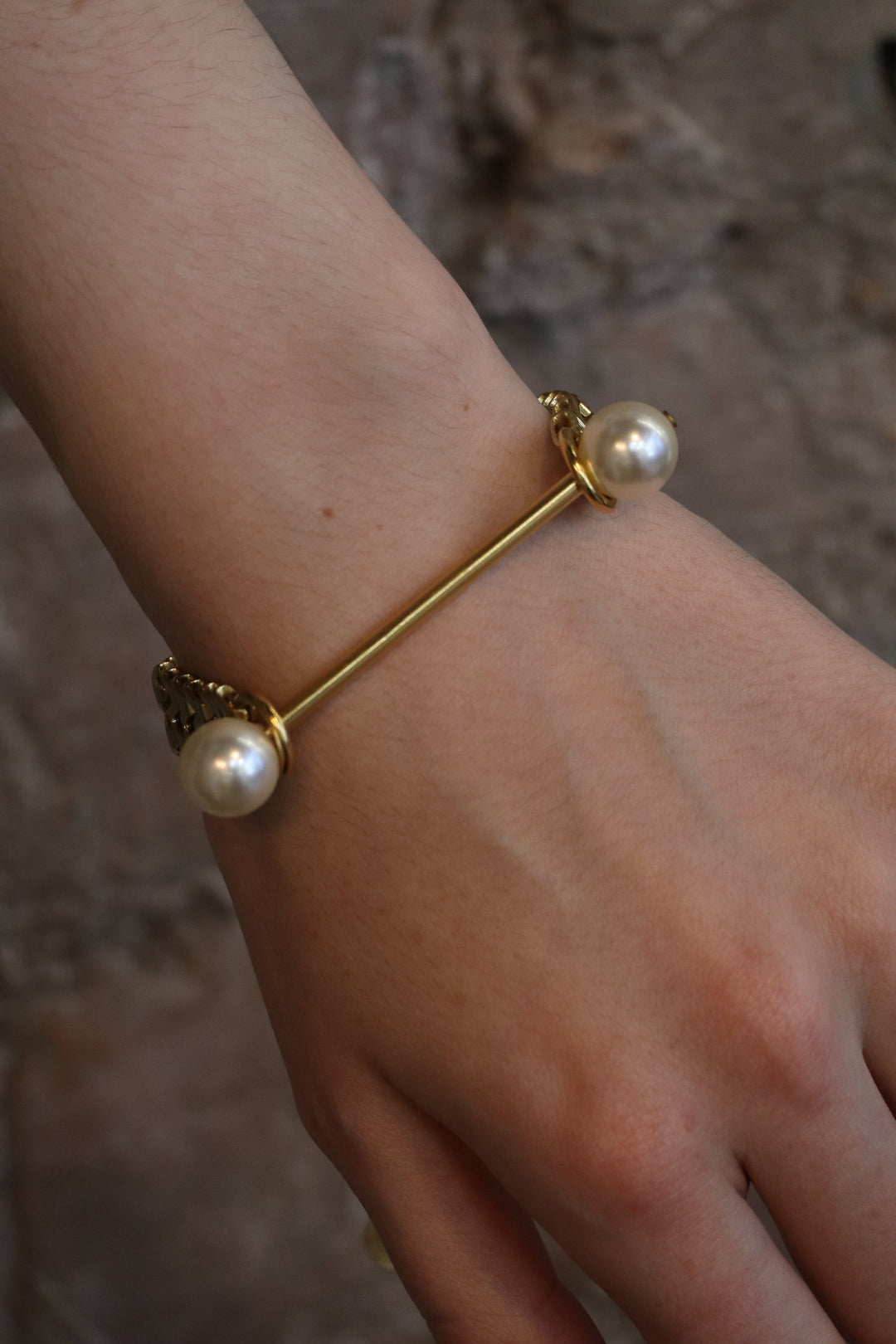 A hand wears the Zubeldia Gold Bar and Double Pearl Chunky Bracelet, featuring a straight gold bar with large white pearls at each end, set against a blurry, textured stone background.