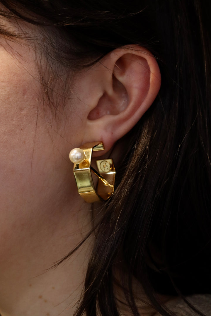 A close-up shows a person wearing Zubeldia Gold Bar and Pearl Hoop Earrings—unique gold-plated hoops with geometric design and single white pearl—framed by their dark hair.