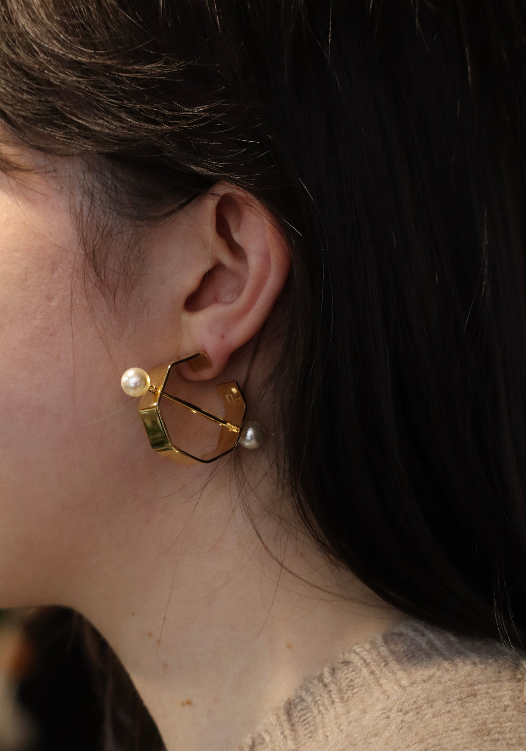 A close-up of a person with long, dark hair and light skin wearing Zubeldia Gold Bar and Pearl Hoop Earrings, featuring a geometric gold plated hoop with two white pearls—one above and one below the lobe.