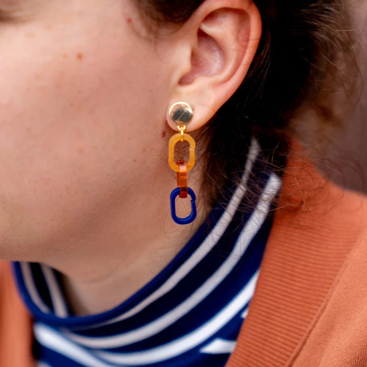 Close-up of a person wearing a beige coat over a blue and white striped top. They have short, curly brown hair and are wearing Studio Nok Nok Gold, Orange & Navy Fire Acrylic Earrings. Crafted meticulously by hand in Belgium, the background is blurred.