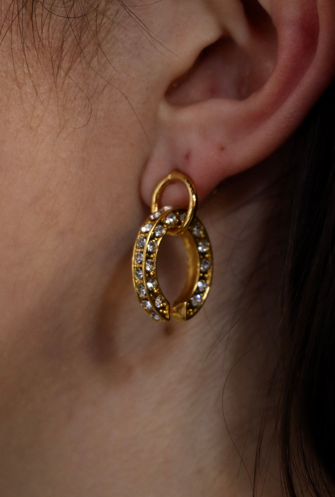 A close-up of a person’s ear wearing Zubeldia Gold Plated Crystal Oval Earrings, featuring a unique overlapping double-hoop design encrusted with sparkling stones for an elegant statement.