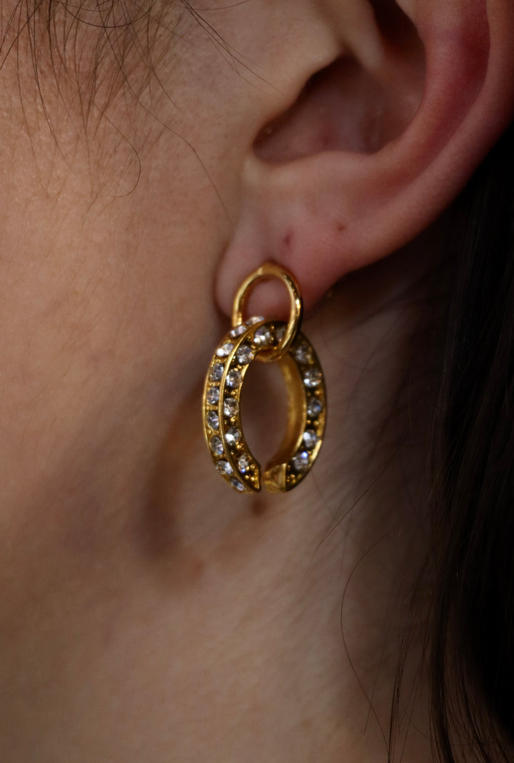 A close-up of a person’s ear wearing Zubeldia Gold Plated Crystal Oval Earrings, featuring a unique overlapping double-hoop design encrusted with sparkling stones for an elegant statement.