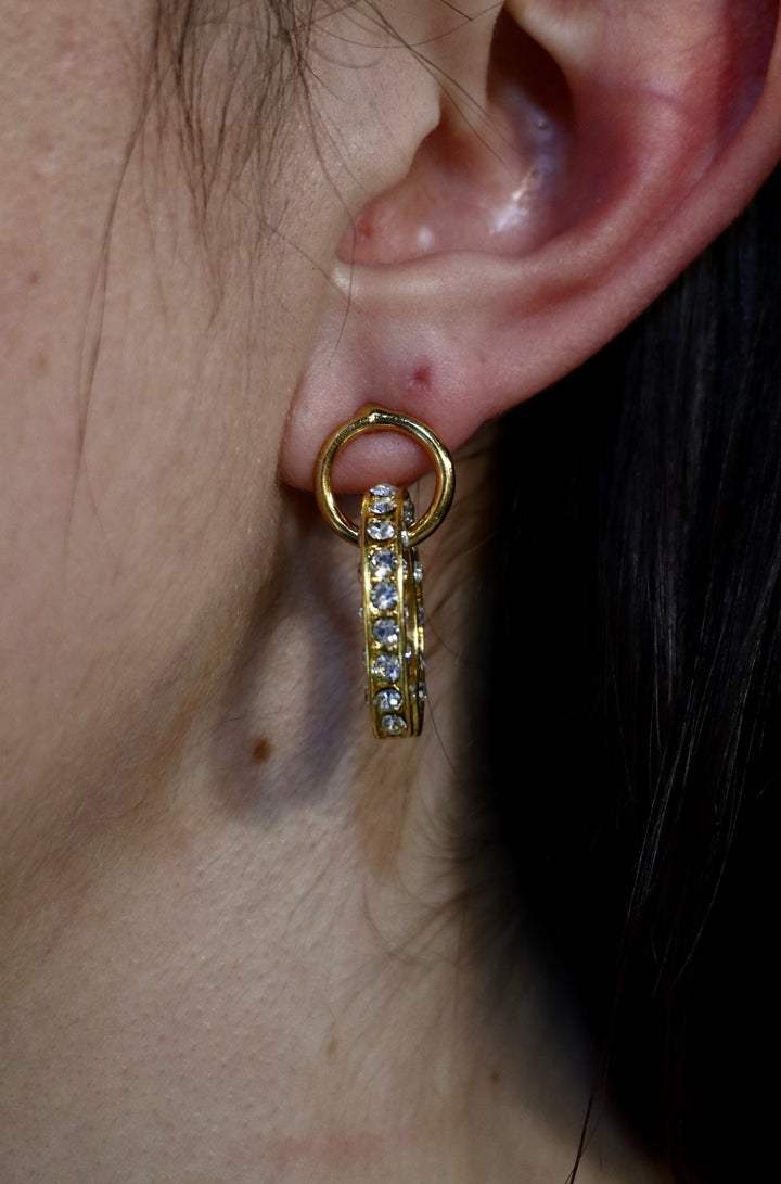 Close-up of a person wearing Zubeldia Gold Plated Crystal Oval Earrings, featuring a row of sparkling crystals in an elegant design. The shiny, decorative earrings are partly visible under the person’s hair.