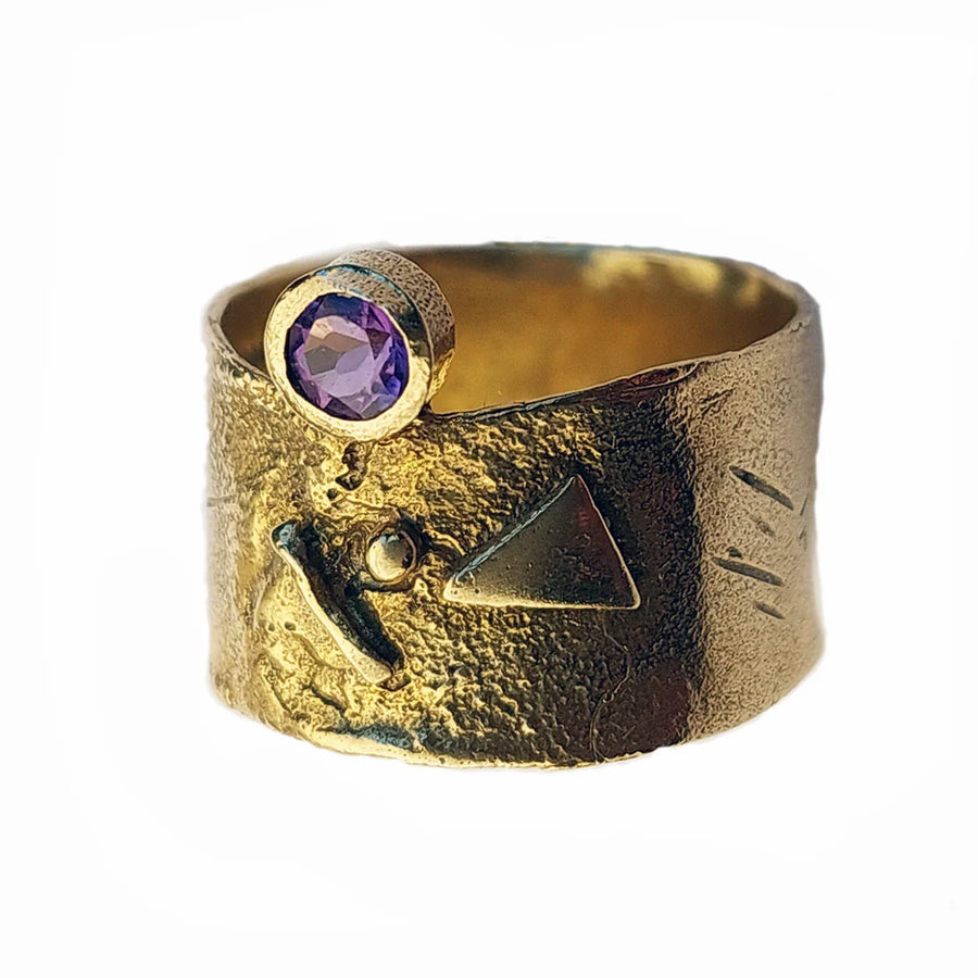 A wide, textured gold Amethyst Guardian Ring Gold by Caroline Stokesberry-lee features a round amethyst gemstone set in a bezel at the top. Engravings on the 18ct yellow gold-plated ring include a triangle and abstract lines, adding an artistic and unique touch to the design.