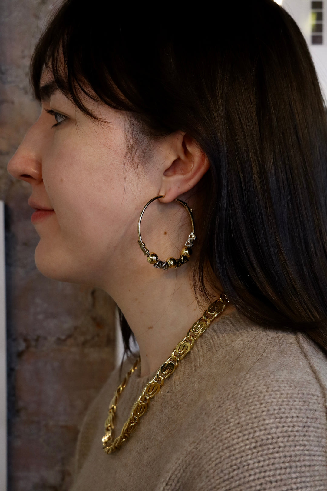 A woman with long dark hair, in a beige sweater and gold chain necklace, wears Zubeldia Gold and Silver Hoop Earrings. She is shown in profile against a brick wall background.