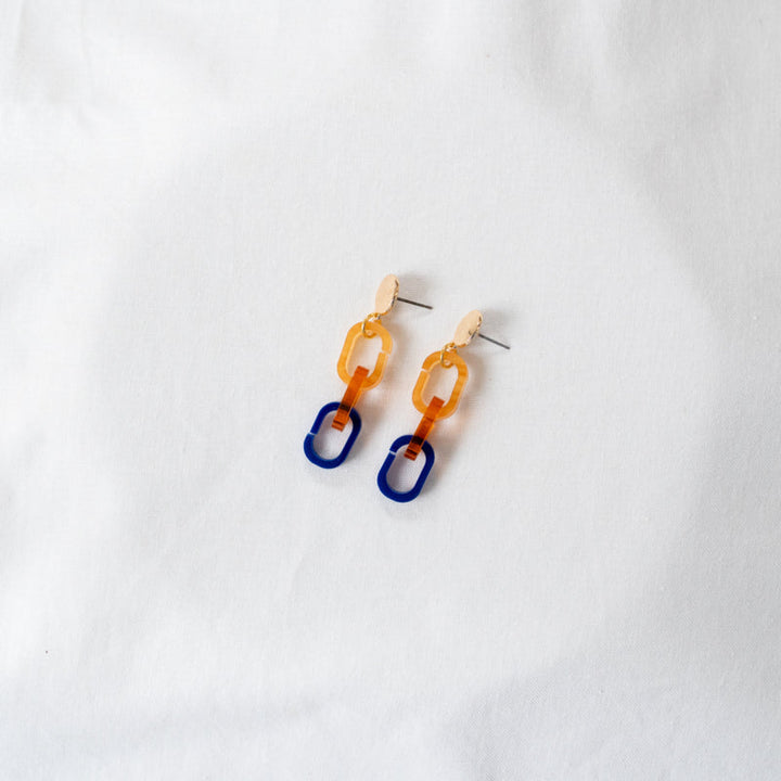 A pair of Gold, Orange & Navy Fire Acrylic Earrings by Studio Nok Nok with geometric linked shapes. Handmade in Belgium, they feature an oval-shaped stud at the top, followed by an amber-colored ovular link, and then a blue rectangular link at the bottom. The gold-plated nickel-free brass design is set against a plain white background.