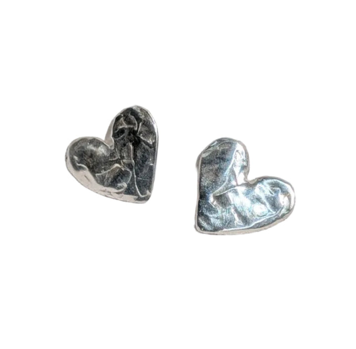 The Gra Silver Heart Stud Earrings by Becca Calmont, featuring a shiny textured design, are displayed on softly folded white fabric under natural light.