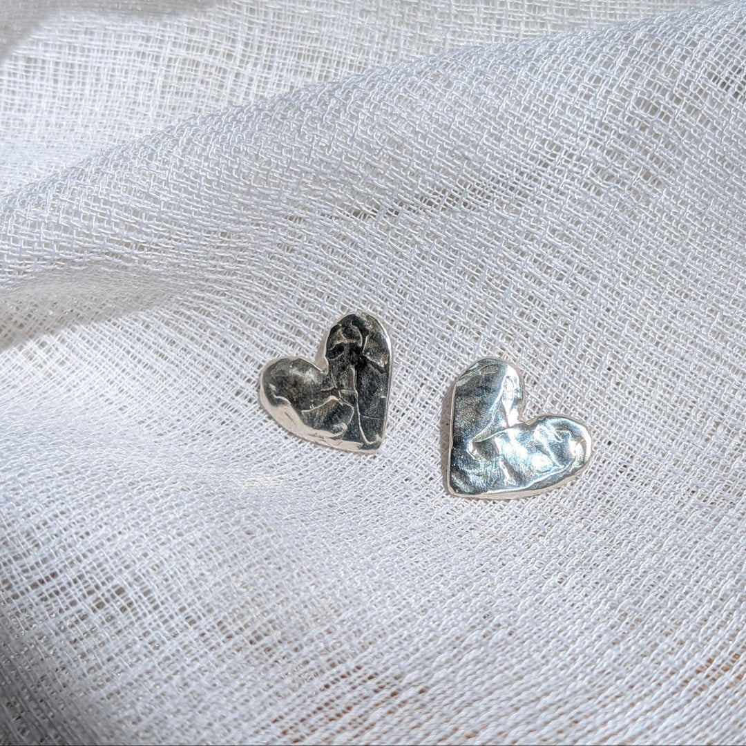Two Gra Silver Heart Stud Earrings by Becca Calmont, handmade from recycled sterling silver, rest on textured white fabric and are softly illuminated by natural light.