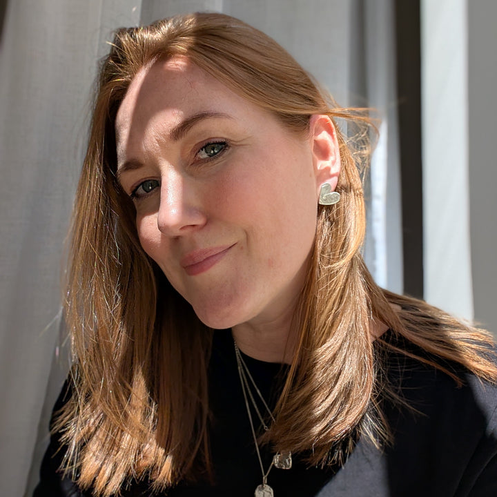 A woman with light brown hair and fair skin smiles softly by a sunlit window with sheer curtains, wearing Becca Calmont's Gra Silver Heart Stud Earrings and layered necklaces. She is dressed in a black top.